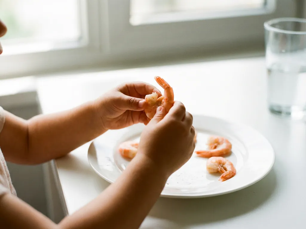 Klein kind houdt voorzichtig een gekookte garnaal vast aan een witte keukentafel, meer garnalen op een bord ernaast