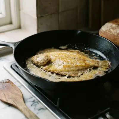 Verse plaice filet sist in zwarte gietijzeren pan met gouden boter, houten spatel op marmeren aanrecht