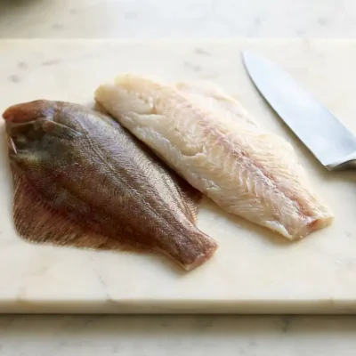 Verse scholfilet en kabeljauwfilet op witte marmeren snijplank met koksmes, natuurlijk daglicht